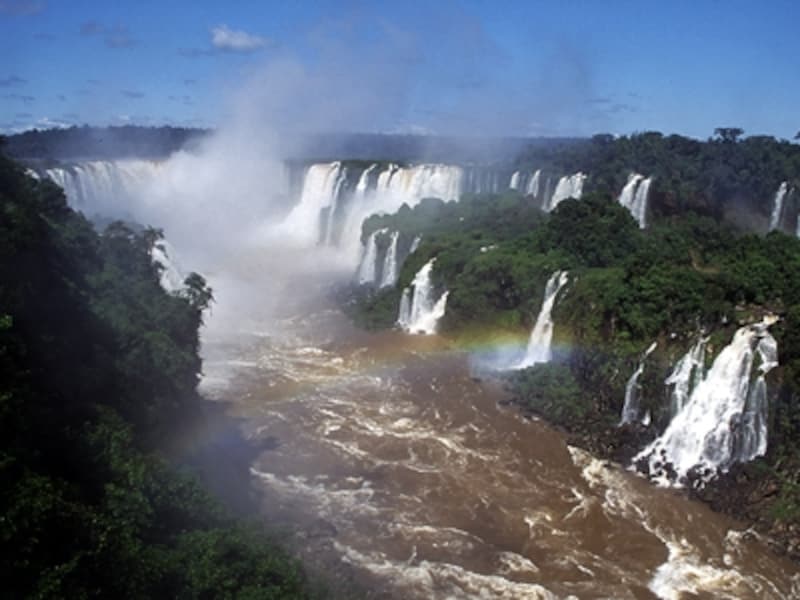 ブラジル側から見たイグアスの滝のハイライト、悪魔の喉笛。酸化鉄からなるラテライトが混ざり込んだ赤い水の流れ、濃い青空に舞い上がるまっ白な水煙、そして7色の虹　©牧哲雄