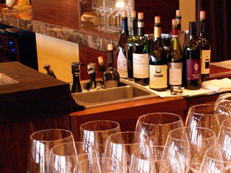 手前に数多く並ぶワイングラス、奥には抜栓してコルクやストッパーでふたをしたワインボトルが林立する