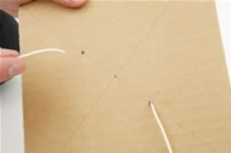3.30cmくらいのタコ糸2本をそれぞれの穴に通します。ダンボール紙をタコ糸の中心にくる位置にあわせて、その両脇にこぶを作り、ダンボール紙が左右にずれないようにします。