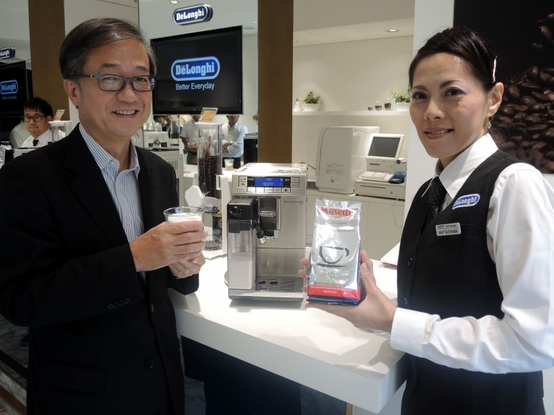 エスプレッソマシンで抽出したカフェラテを手に笑顔の杉本氏（左）