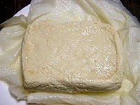 豆腐はキッチンペーパーなどにくるみ、電子レンジで3分加熱します。その後レンジから取り出し、重しをして、元の厚みの半分近くになるまでよく水切りします。<br />