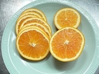 オレンジは50℃くらいの湯に10分ほど浸けておき、湯の中で指先でごしごしこすって洗ったあと、水で洗い流して表面についたワックスを落とす。オレンジの中ほどの部分を、8枚ほど薄くスライスする。<br />