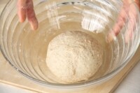 表面を張らせるように、生地を裏でつまみとめて丸め、ボウルに入れる。固くしぼったぬれ布巾をかけて、オーブンの発酵機能を利用するなら30℃で50～60分、25℃くらいの室温で1時間～1時間半、生地がひと回り大きくなるまで、発酵させる。<br />