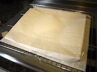 ケーキクーラーの上にオーブンペーパーを敷き、その上にひっくり返した生地をおき、乾かないようにわら半紙をつけたまま、あら熱をとります<br />