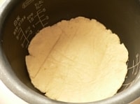 内釜の底の部分を、分量外の油をつけたペーパーでふく。（4）の生地を、グラニュー糖がついてる方を下にして入れ、端の部分を釜に押しつけて貼りつける。<br />