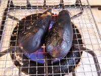 コンロに網をのせるか、魚焼きグリルで<br />
茄子を焼く。ときどき返しながら、<br />
まんべんなく火が通るように焼いて<br />
冷水で冷ましながら、皮をきれいに除いて<br />
へたを落とし、一口大に切る。<br />