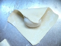 パイシートを麺棒で正方形に伸ばし、真ん中に大福を置いて、上下左右のパイを真ん中に寄せて重ねて包む。重なる部分にはドリール（卵液）をぬって、指で押さえてくっつける。<br />